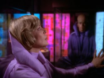 Barbara is a a glass tank in a brightly and colourfully lit room. She is wearing elaborate pink satin sci-fi robes and looking off into the\ distance. In the background is an alien man with a massive domed head.