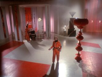 Barbara stands in the middle of a massive high-ceilinged room with stylish décor in red and white. She is wearing her spacesuit with no helmet. Her arms are held out pleadingly.