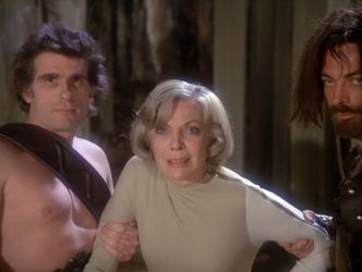 Barbara is being held by the arms by two hairy men. She looks distressed.