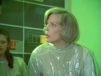 Barbara is in a quarantines operating room with green-lit walls. She's wearing her now customary cling film scrubs. She looks up at something off camera, startled.