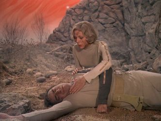 Barbara is on a strange planet with a cyclorama sky painted in soft red swirls, with dead vegetation and a fibreglass cliff in the background. She is covered in dirt. Before her, lying on the ground, is Koenig's body. She reaches out her hand to touch it gently.