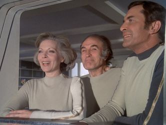 Barbara, Victor and John look out of a Main Mission window onto the moon's surface. Their faces  are bathed in sunlight. Barbara is smiling, with the wind blowing through her hair.